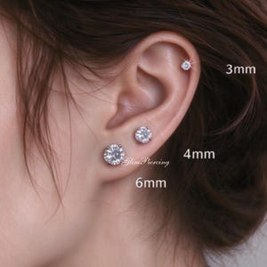 Könnte beinhalten: Drei runde, klare Ohrstecker mit Krappenfassung, die in einem Ohr getragen werden. Die Ohrringe sind mit den Größen 3mm, 4mm und 6mm neben dem Text GlimPiercing beschriftet. Jeder Stecker verfügt über einen facettierten Stein im Brillantschliff.