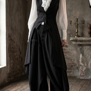 Könnte beinhalten: Eine weiße Langarmbluse mit hohem Rüschenkragen und einer Kamee-Brosche, getragen unter einer schwarzen ärmellosen Weste mit zwei Knöpfen. Das Outfit umfasst eine schwarze weite Hose mit seitlichen Metallringen und einem asymmetrischen Saum.
