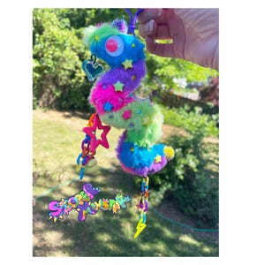 Puede incluir: Un llavero colorido en forma de gusano de peluche con un ojo grande, estrellas y colgantes. El gusano es una mezcla de pelo azul, verde, morado y rosa. El llavero incluye un corazón, una estrella y un rayo, y el texto "Spoopy Worms".