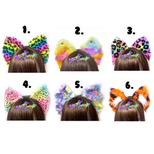 Puede incluir: Seis pares de orejas de gato de piel sintética en varios patrones y colores, incluyendo estampado de leopardo arcoíris, tie-dye pastel y rayas de tigre naranja. Cada par incluye un dije decorativo con el texto "Stoked Monster".