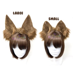 Op de afbeelding: Twee paar bruine nep bont dierenoren, een paar met het label "LARGE" en het andere paar met het label "SMALL". De oren worden gedragen op een bruin pruik op een mannequinhoofd.