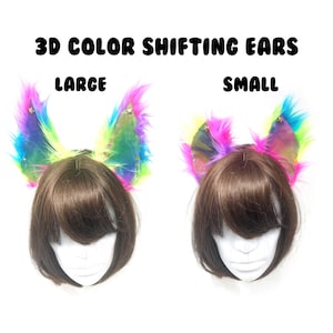 Puede incluir: Dos pares de orejas 3D que cambian de color, etiquetadas como "LARGE" y "SMALL". Las orejas están unidas a cabezas de maniquí con cabello castaño. Las orejas son una mezcla de colores del arcoíris, incluyendo rosa, amarillo, azul y verde.