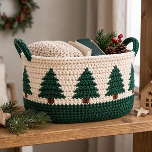 May include: A round, cream-colored crocheted basket featuring a repeating pattern of dark green pine trees with brown trunks. The basket has two dark green handles and a solid dark green base, filled with a textured beige fabric and greenery.