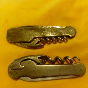 May include: Two vintage metal pocket corkscrews with integrated bottle openers. The top corkscrew features the word EINA stamped on the handle. Both tools have a folding design with a metallic finish showing signs of age and wear.