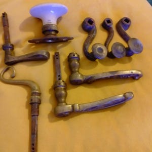 Peut inclure: Une collection de quincaillerie de porte vintage comprenant un bouton en porcelaine blanche avec une base en laiton, trois petits crochets en laiton à extrémités circulaires et plusieurs poignées de porte en laiton avec des tiges carrées.