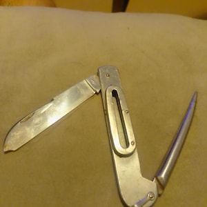 Peut inclure: Un couteau de poche multifonction argenté doté d'une lame pliante d'un côté et d'un épissoir pointu de l'autre. Le manche central présente une découpe rectangulaire et une petite boucle métallique à la base, avec le mot Japan gravé sur la lame.