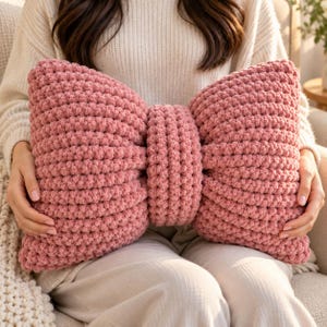 May include: A dusty rose pink bow-shaped throw pillow crafted from thick, chunky knit yarn. The pillow features a central knotted section with two large, voluminous loops on either side, creating a distinct bow silhouette with a textured surface.