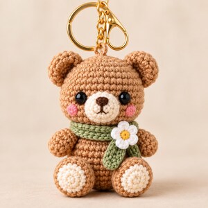 May include: A brown crochet bear keychain featuring a light beige muzzle, black plastic eyes, and pink cheeks. The bear wears a sage green scarf adorned with a small white flower with a yellow center. It is attached to a gold metal keyring.