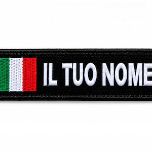Puede incluir: Un parche rectangular bordado en negro con la bandera italiana en el lado izquierdo y el texto blanco IL TUO NOME en una fuente sans-serif en negrita a la derecha. El parche tiene un borde cosido en negro y una superficie de tela texturizada.