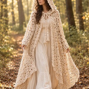 May include: A floor-length, cream-coloured crochet hooded cloak featuring an intricate openwork pattern and a front tie closure with long tassels. The garment is worn over a matching cream-coloured, tiered, long-sleeved maxi dress.