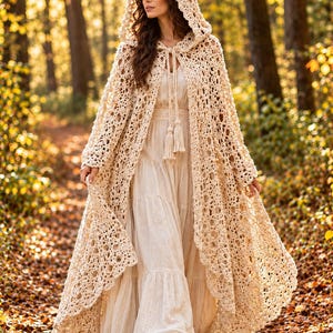 May include: A floor-length cream-colored crochet hooded cloak featuring an intricate openwork pattern and a front tie closure with long tassels, worn over a matching cream-colored tiered maxi dress with a simple bodice and long skirt.
