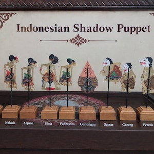 Puede incluir: Una colección de diez marionetas de sombra indonesias exhibidas en soportes de madera individuales. Las figuras incluyen a Sadewa, Nakula, Arjuna, Bima, Yudhistira, Gunungan, Semar, Gareng, Petruk y Bagong, con detalles pintados.