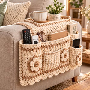 May include: A beige and cream crochet sofa armrest organizer featuring multiple pockets, scalloped edges, and decorative flower motifs. It holds a remote control, glasses, a notebook, a smartphone, a ceramic mug, and a small potted plant.