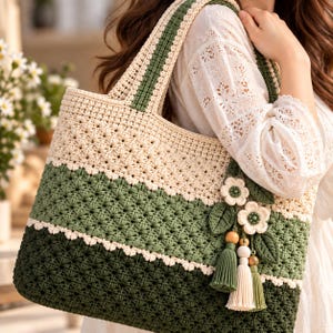 May include: A crochet tote bag featuring a color-blocked design in cream, sage green, and dark forest green. The bag has a textured star-like stitch pattern and is decorated with two crocheted flowers, green leaves, and three hanging tassels.