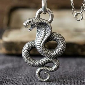 Puede incluir: Un collar de metal plateado con un colgante de cobra detallado, con la boca abierta y escamas texturizadas. La serpiente está enroscada en forma circular y cuelga de una fina cadena plateada con un cierre de resorte redondo.