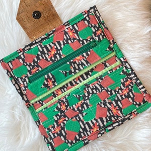 May include: A rectangular fabric wallet featuring a black background with a repeating pattern of green potted cacti and small orange flowers. The wallet includes a green zippered compartment, multiple card slots, and a cork flap with a metal snap.