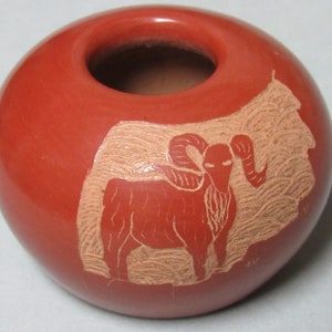 May include: A red clay pot with a carved design of a ram with large horns. The ram is carved in a lighter shade of clay, creating a contrast against the red background.