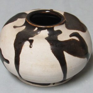 May include: A white ceramic vase with brown glaze dripping down the sides. The vase has a round body and a wide opening.