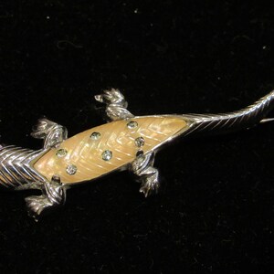 May include: A silver and cream colored lizard brooch with rhinestones. The lizard is detailed with a textured body and a long tail.