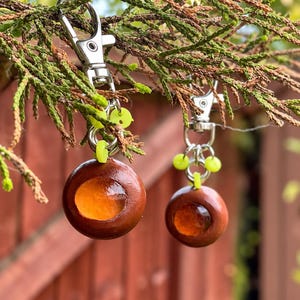 May include: Two round, polished brown wooden keychains featuring a central, translucent amber-coloured resin inlay. Each keychain is attached to a silver-toned metal lobster clasp by a series of small, bright neon yellow plastic rings.
