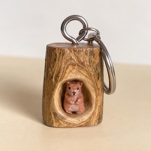 May include: A wooden keyring featuring a cylindrical log segment with a carved hollow centre. Inside the hollow, a small, light brown, hand-painted animal figure is visible. A silver-toned metal eye screw and a split ring are attached to the top.