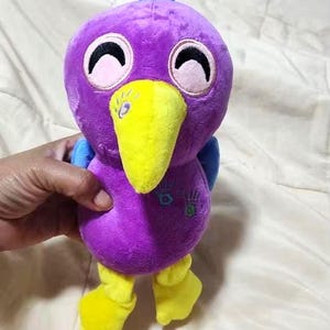 May include: A plush toy shaped like a bird with purple fur, a bright yellow beak, and yellow feet. The bird has blue wings, a blue tuft on its head, and closed eyes. There are small embroidered handprint designs on the chest area of the plush.