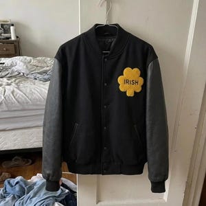 Peut inclure: Une veste universitaire noire avec des manches longues noires, un col côtelé et une fermeture à boutons sur le devant. Un écusson jaune texturé en forme de trèfle est sur la poitrine gauche avec le mot IRISH brodé en lettres noires.