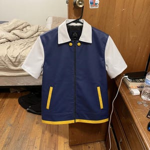 Könnte beinhalten: Eine kurzärmelige Jacke mit marineblauem Rumpf, weißen Ärmeln und weißem Kragen. Das Kleidungsstück verfügt über einen durchgehenden Frontreißverschluss, zwei gelbe vertikale Taschenakzente, einen gelben Saum und zwei gelbe runde Knöpfe am Kragen.