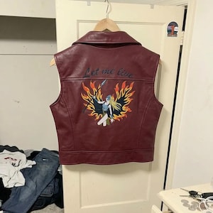 May include: A sleeveless burgundy vest with a wide collar, hanging on a wooden hanger. The back features an embroidered graphic of a figure with long blonde hair holding a spear, surrounded by orange and yellow flames, with the text Let me live.