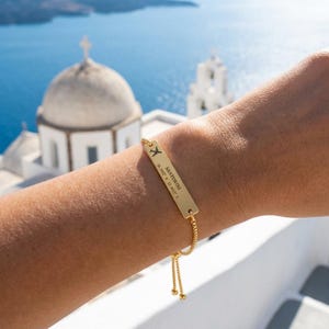 Puede incluir: Una pulsera de metal dorado con una placa rectangular grabada con un icono de avión y el texto SANTORINI 36.3932 N 25.4615 E. La pulsera cuenta con una cadena fina con un mecanismo deslizante ajustable y dos pequeñas cuentas en los extremos.