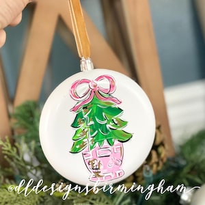 May include: White round Christmas ornament with a hand-painted design of a green Christmas tree with a pink bow on top, in a pink pot with gold accents. The ornament has a gold string for hanging.