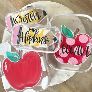 May include: Three clear plastic containers featuring hand-painted designs. Two containers display red apples with green leaves and white polka dots. One rectangular container features a yellow pencil design with the text Kristen and Mrs. Hopkins.