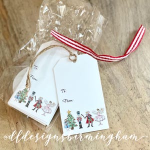 May include: A set of white gift tags with a Nutcracker theme. The tags feature a watercolor illustration of a Christmas tree, a Nutcracker, and a ballerina. The tags have the words "To:" and "From:" printed on them.
