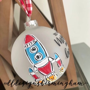 May include: A white frosted glass ornament with a hand-painted rocket ship design. The rocket ship is red, blue, and white with black outlines. The ornament also has the text "To the Moon and Back" written in black.