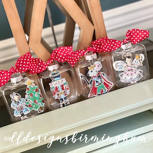 May include: Four clear glass ornaments with red and white polka dot ribbon. Each ornament features a different character from the Nutcracker ballet: a Christmas tree, a nutcracker soldier, a mouse king, and a sugar plum fairy.