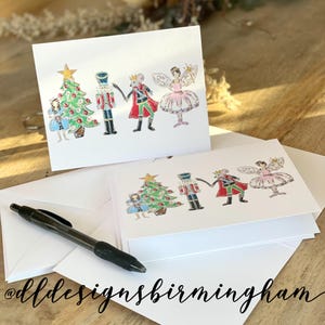 May include: A set of four Christmas cards featuring a whimsical illustration of the Nutcracker, a mouse king, a sugar plum fairy, and a Christmas tree. The cards are white with a subtle shimmer and are perfect for sending holiday greetings.