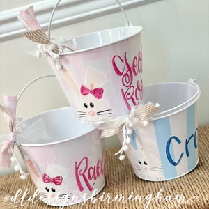 May include: Three Easter buckets with white handles, each painted with a bunny design and a name in cursive. The buckets are pink and blue, with striped and brushstroke patterns. Each bucket has a decorative bow.