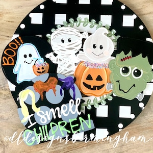 May include: A black and white checkered background with a wreath of green leaves.  A white ghost with a pumpkin bucket, a mummy, a ghost with a pumpkin, a pumpkin with a jack-o-lantern face, and a green Frankenstein monster are arranged around the wreath.  The text "BOO!" is on the left side of the image.  The text "I smell children" is on the bottom of the image.
