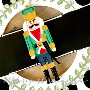 May include: A painted wooden nutcracker figurine with a green and gold uniform, a red sash, and a black mustache. The nutcracker is standing on a white surface with a black background.