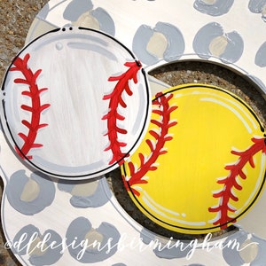 May include: Two painted wooden baseballs, one white with red stitching and one yellow with red stitching. The baseballs are on a white background with a gray leopard print pattern.