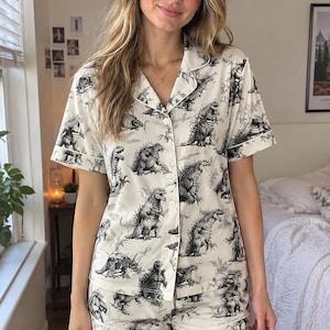 May include: A cream-colored pajama set featuring a short-sleeved button-down shirt and matching shorts. The fabric is decorated with a repeating black and white graphic print of Godzilla monsters and foliage throughout the entire garment.