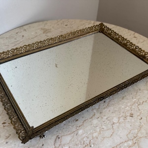 May include: A rectangular vanity tray featuring a mirrored base with a speckled, aged patina. The tray is enclosed by an ornate, perforated gold-tone metal border with intricate filigree detailing and small feet at the corners.