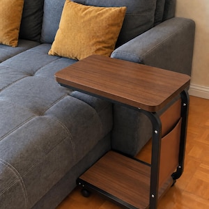 Op de afbeelding: Een C-vormige bijzettafel met een blad en onderste plank in donkere houtnerf, ondersteund door een zwart metalen frame. De zijkant is voorzien van een bruin opbergvak en de basis is uitgerust met vier kleine zwarte zwenkwielen voor mobiliteit.