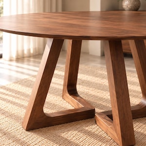 Puede incluir: Una mesa de comedor redonda hecha de madera de color marrón oscuro con una superficie circular lisa. La mesa se apoya en una base única que consta de tres patas de madera anguladas e interconectadas que forman una estructura geométrica.