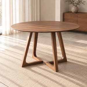 Puede incluir: Una mesa de comedor redonda fabricada en madera de tono marrón medio con una superficie circular lisa. La mesa presenta un diseño de base único que consta de tres patas de madera anguladas e interconectadas para soporte.