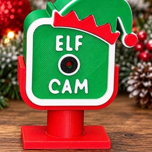 May include: A green and red plastic decorative sign shaped like a camera with a green elf hat on top. The sign features the white text "ELF CAM" and a black circular lens in the centre. It is mounted on a red cylindrical post and a square base.