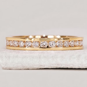 Könnte beinhalten: Ein Ring aus Gelbgold mit einer Kanalfassung, in der eine durchgehende Reihe runder, klarer, facettierter Steine in der Mitte eingelassen ist. Das Metall hat eine polierte, reflektierende Oberfläche und die Steine sind linear angeordnet.