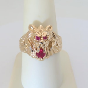 May include: A gold-toned ring featuring a lion's head design. The lion's eyes and mouth are accented with vibrant red gemstones. The ring is displayed on a white stand, showcasing its intricate details and craftsmanship.