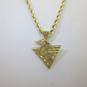Puede incluir: Un collar de cadena de oro con un colgante que presenta un dise&ntilde;o de p&aacute;jaro estilizado. El colgante tiene forma triangular y una superficie texturizada.