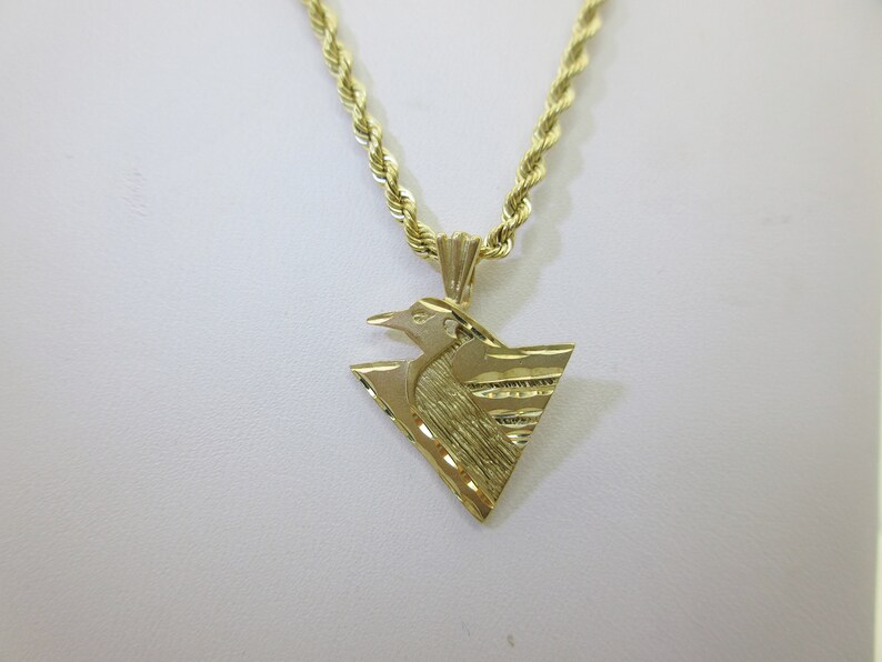 Puede incluir: Un collar de cadena de oro con un colgante que presenta un dise&ntilde;o de p&aacute;jaro estilizado. El colgante tiene forma triangular y una superficie texturizada.